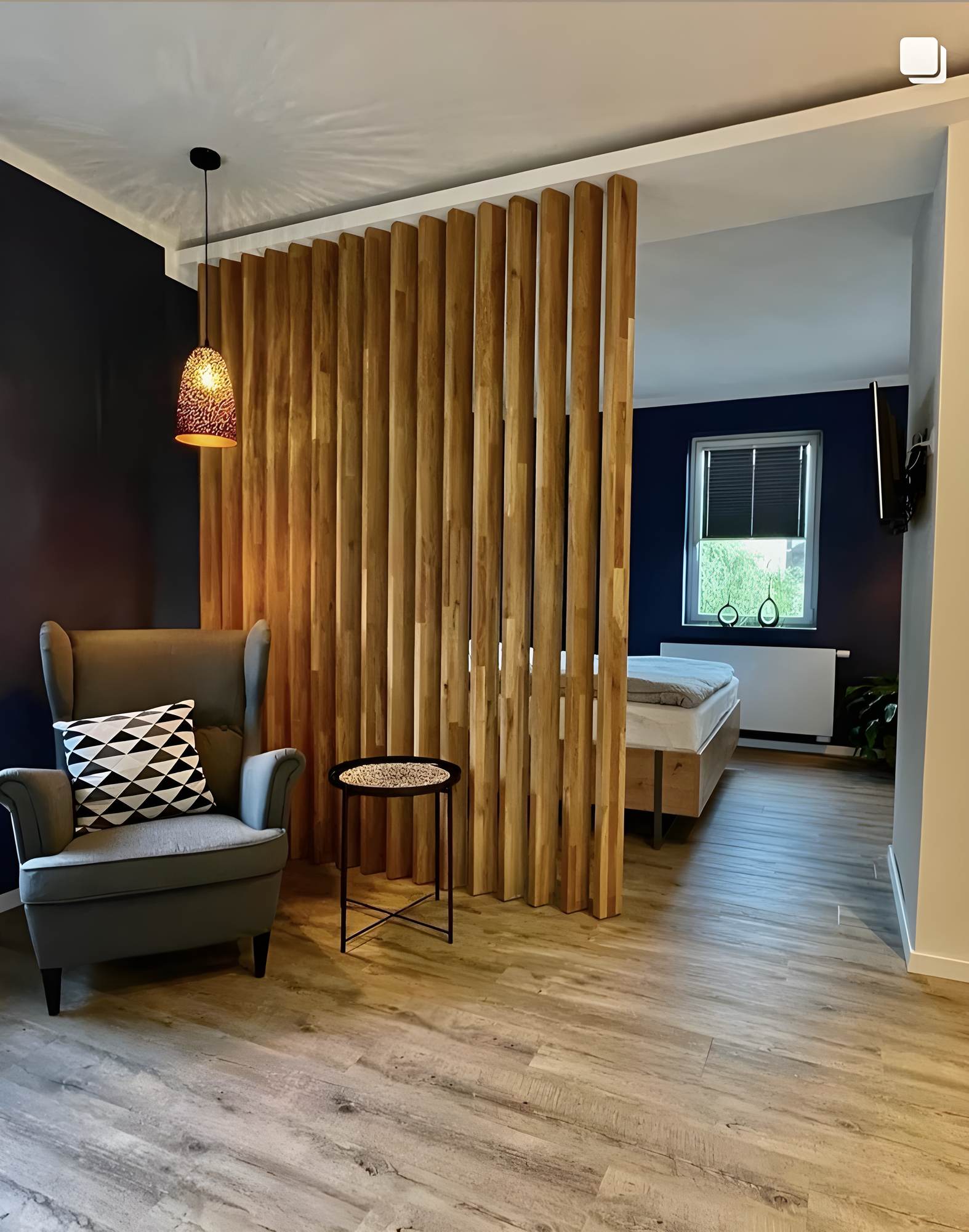 Schlafzimmer mit Raumteiler aus freistehenden Holzlamellen, Sessel und warmer Pendelleuchte